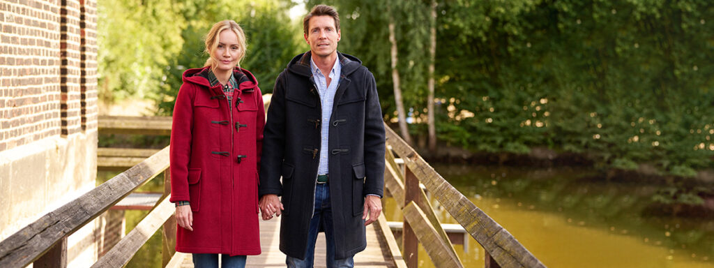 Lady in a red duffle coat and gentleman in a dark blue duffle coat, holding hands on a bridge